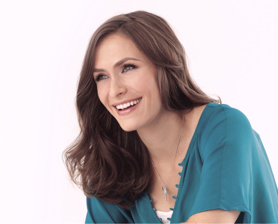A woman with long brown hair laughs broadly in a teal blouse and a necklace.