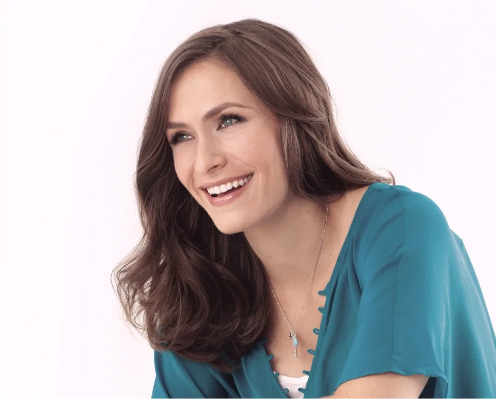 A woman with long brown hair laughs broadly in a teal blouse and a necklace.
