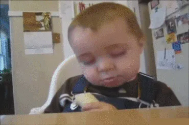 A sleepy baby with closed eyes chews a snack while seated at a table near a refrigerator.