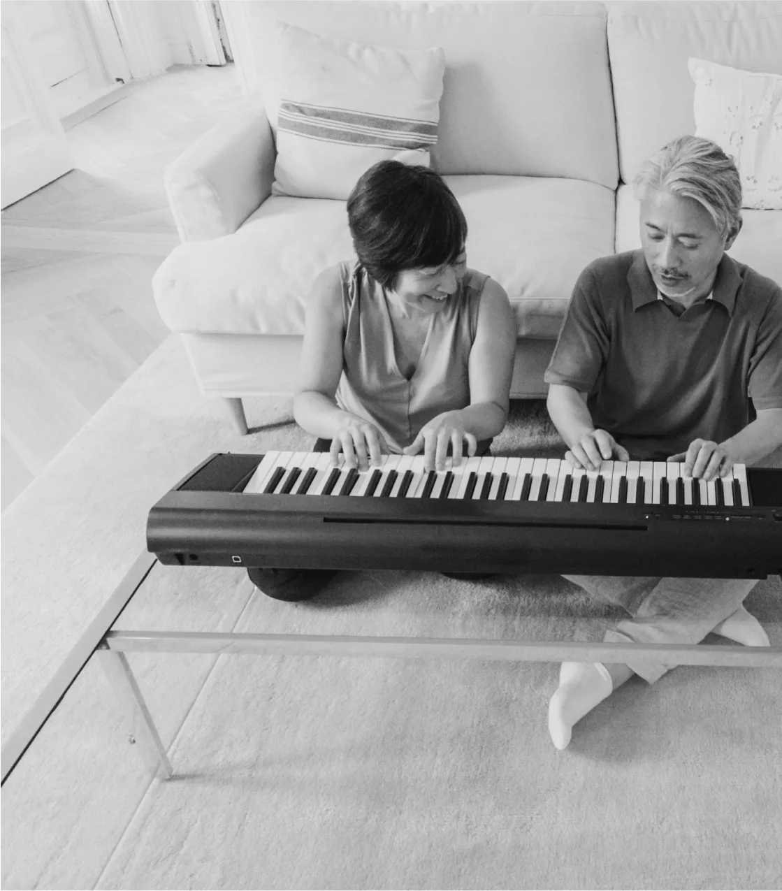 Image-2.jpg An older couple sit on the floor playing an electric keyboard together in a bright living room, in black and white.