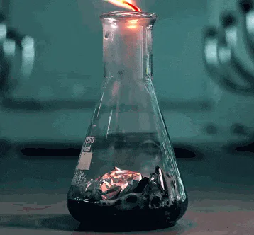 A glass flask on a lab bench produces a bright green flame that grows and flickers upward.