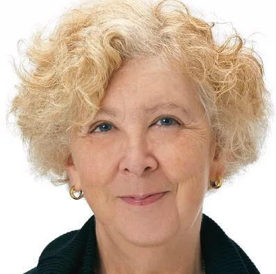 Screenshot+2024-06-25+085955.png An older woman with curly gray-blonde hair and hoop earrings wears a black top against a white background, with a neutral expression.
