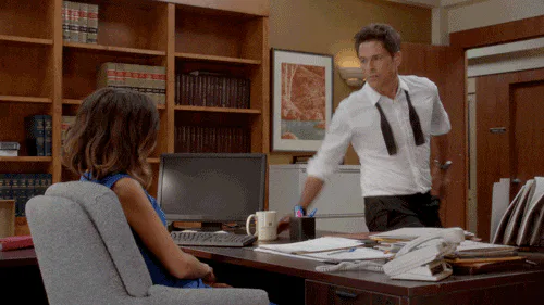 A man reaches across a desk, sending papers flying, as a seated woman looks on.