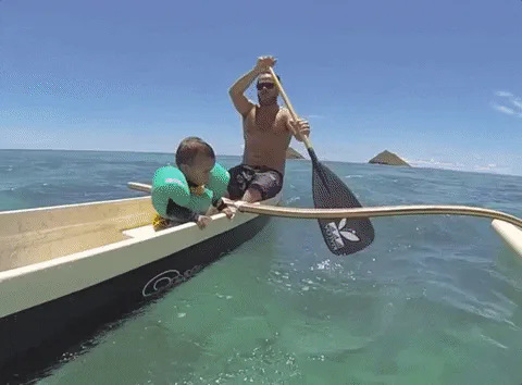 A man and a young boy in a life vest wrestle and splash off the side of a canoe in the ocean.