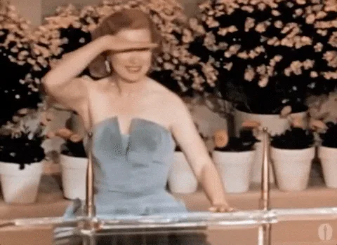 A woman in a strapless blue gown shields her eyes with one hand while seated among potted flowers on a vintage show.