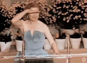 looking+where+are+you+GIF+by+The+Academy+Awards.gif A woman in a strapless blue gown shields her eyes with one hand while seated among potted flowers on a vintage show.