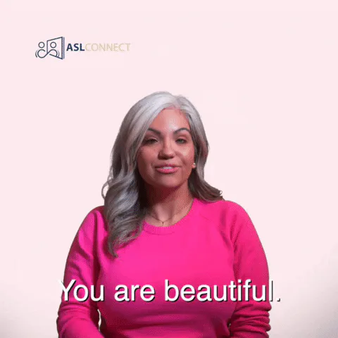 A woman with gray hair signs 'You are beautiful' in American Sign Language against a pink background.