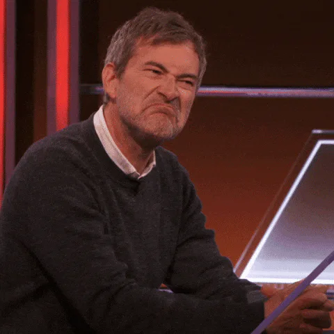 A gray haired man in a black sweater frowns and gestures with both hands while seated at a desk.
