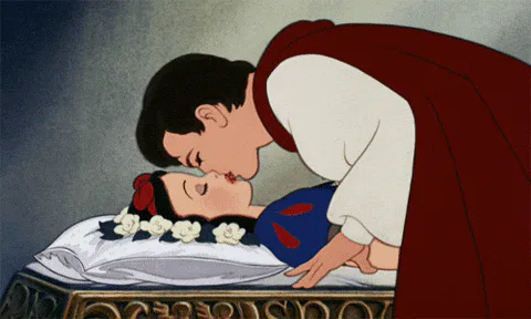 An animated prince leans down to kiss a sleeping princess laid out on a stone bier surrounded by flowers.