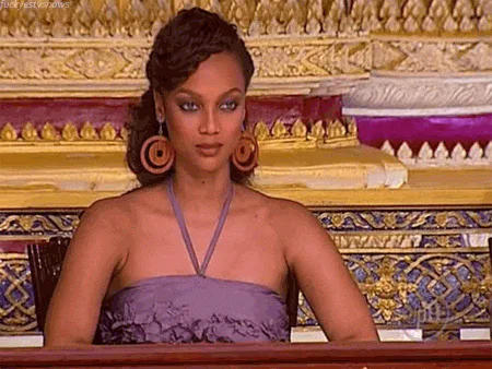 A woman in a purple gown with large earrings sits regally in front of an ornate gold backdrop.
