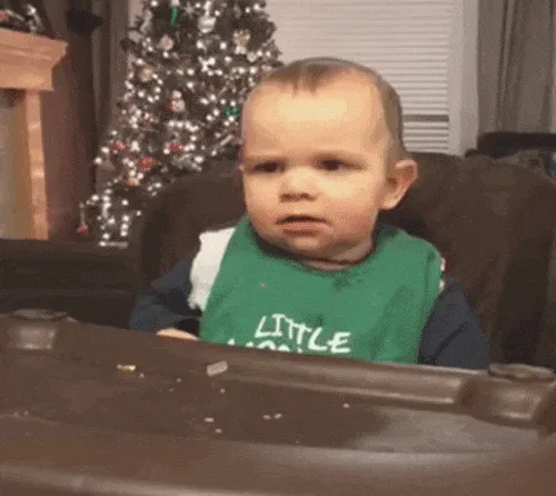 A baby in a green bib sits in a high chair by a Christmas tree and raises one finger.