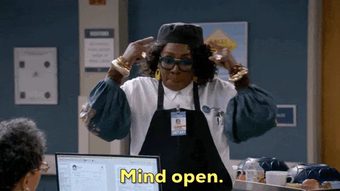 A woman in a black cap and apron raises her arms and points while talking and says 'Mind open.'