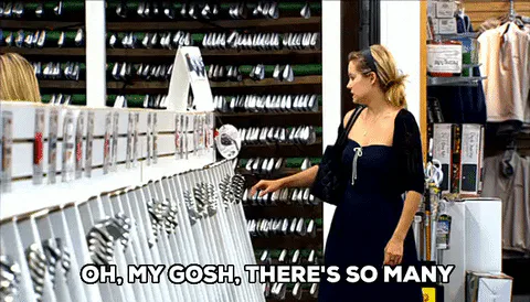 A woman in a black dress browses a long store aisle of tools and says 'Oh my gosh, there's so many.'