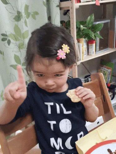 A toddler girl with a flower hair clip holds a snack and points sternly, wearing a shirt reading 'Til to moon.'