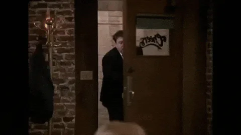 A man in a suit walks through a doorway into a bar, then the scene cuts to a bartender and seated patrons at the bar counter.