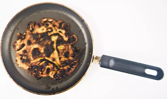 A badly burnt, misshapen pancake sits in a black nonstick frying pan.