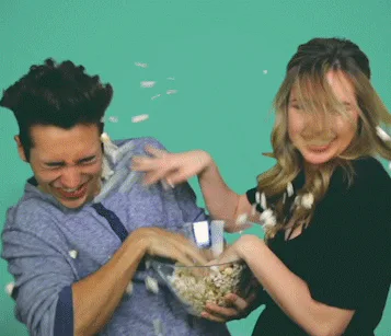 A man and woman on a green background laughingly throw popcorn and toss it into each other's hair.