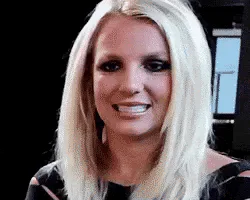 A blonde woman with dramatic eye makeup smiles and laughs nervously at the camera.