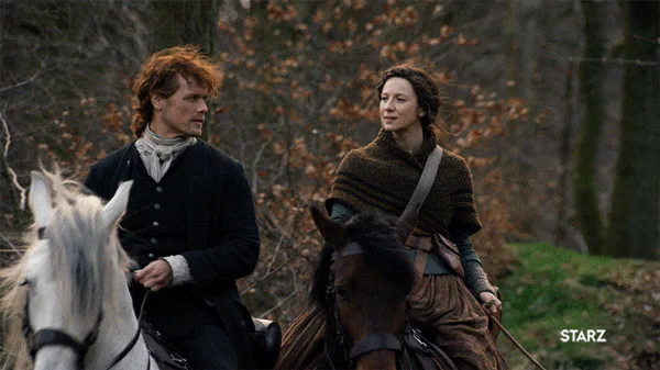 A red-haired man and a brown-haired woman ride horses side by side through an autumn forest.