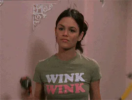 A woman in a shirt reading 'Wink Wink' holds a power drill and a hammer, looking puzzled.