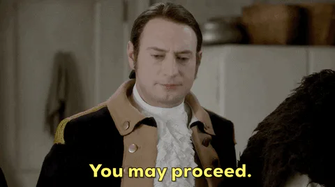 A man in an 18th century coat and ruffled collar says 'You may proceed.'