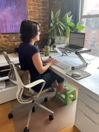 biglaurieposture.jpeg Laurie Gerber sits at a standing desk, typing with a laptop, plant and green drink nearby.