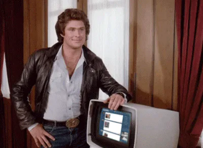 A man in a leather jacket leans against a glowing computer console and smiles.