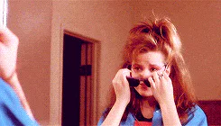A teenage girl with big hair adjusts black sunglasses while looking in a mirror, wearing a blue jacket.