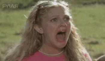 A young blond woman in a pink top screams open mouthed outdoors, with sunglasses pushed up on her head.