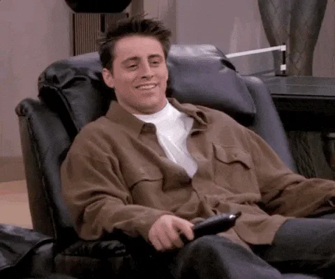 A man in a brown jacket laughs while relaxing in a leather recliner, holding a remote control.