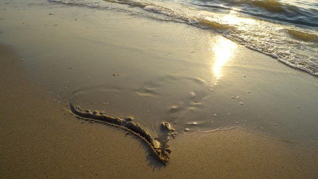 A heart shape drawn in wet sand on a beach catches the sunset's golden light.