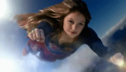 A woman in a superhero costume flies through clouds with her fist forward, hair streaming back.