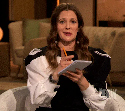 A woman with wavy brown hair sits on a talk show set holding a notepad and pencil while talking.