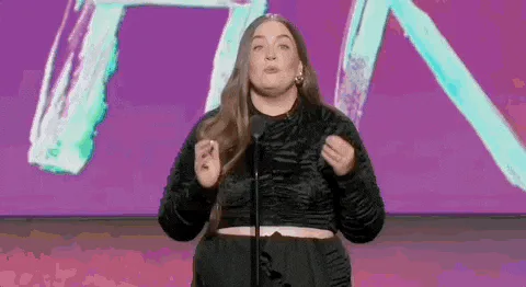 A woman on a colorful stage says 'We love it. We do it. We regret it.'