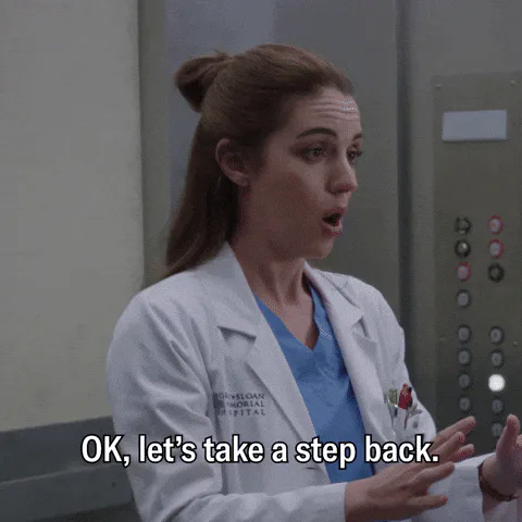 A woman in a white coat and scrubs gestures by an elevator and says 'OK, let's take a step back.'
