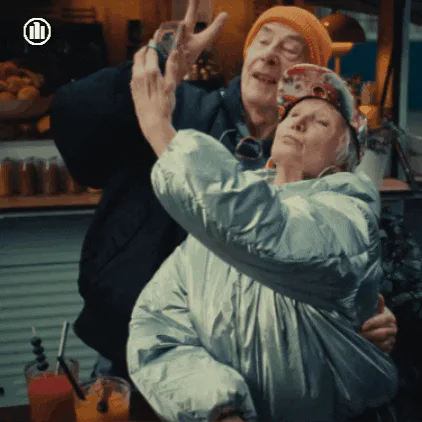 An older couple pose for a selfie together at an outdoor bar counter, smiling.