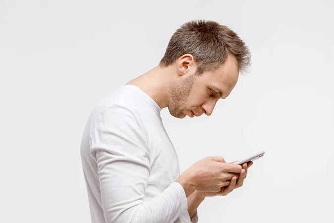 pasted+image+0+(1).png A man in a white shirt hunches forward, craning his neck to look at his phone.
