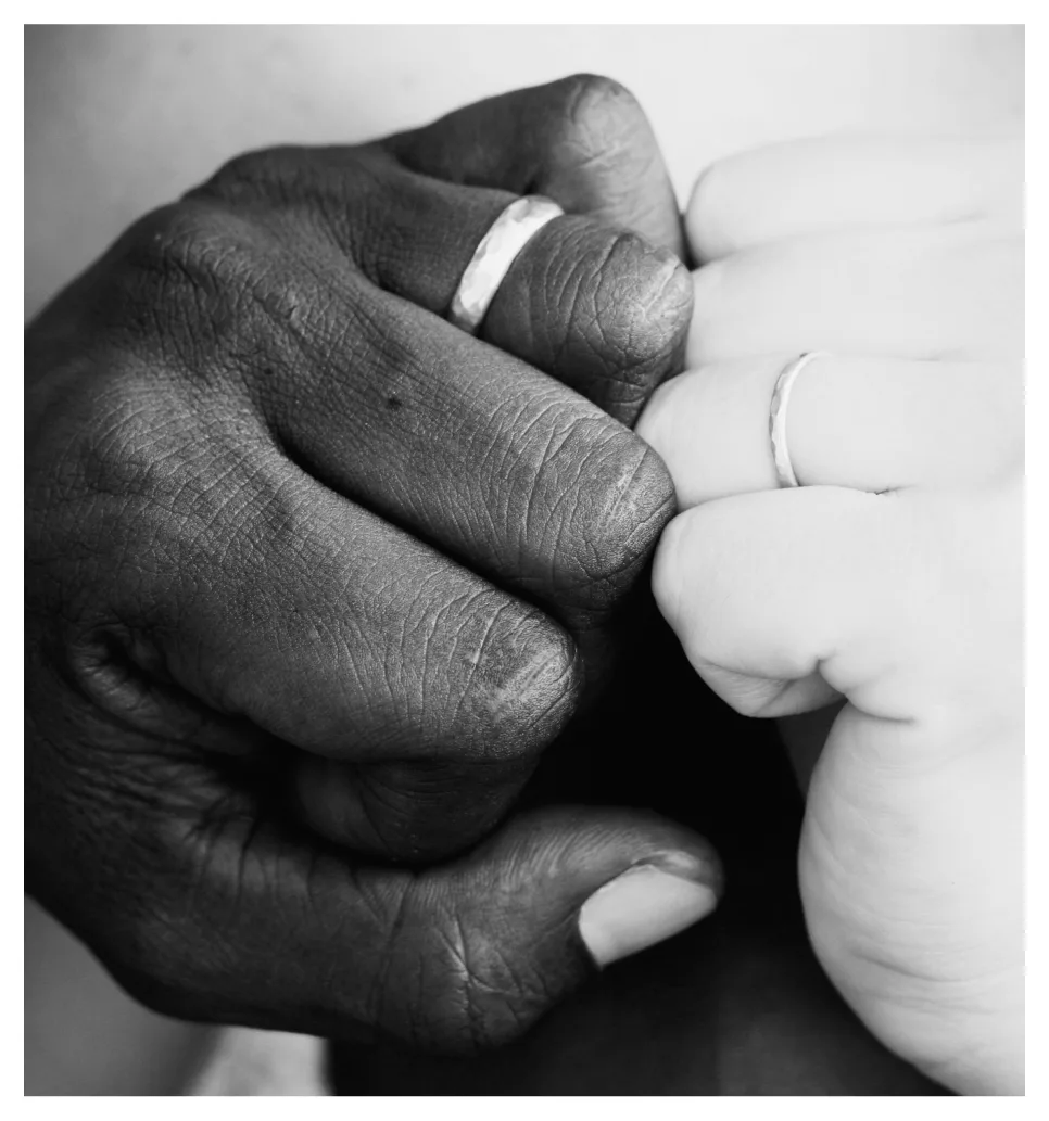 A black and white close-up of two clasped hands, each wearing a simple wedding band.