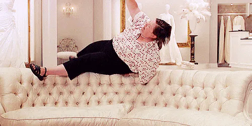 A woman in a floral top tumbles backward onto a cream sofa in a bridal shop.